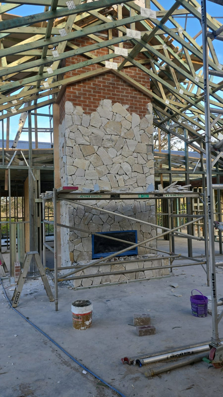 A Cresta Beige Stone Cladding fireplace with split-face texture and a brick chimney is being built inside a partially constructed house, surrounded by wooden framing, scaffolding, and tools scattered on the concrete floor. — Stone Cladding