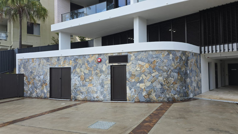 A building exterior features Verona Stone Cladding with free style walling, two dark doors, a central vented door, and wet natural bluestone paving bordered by stone. Palm trees and balconies appear in the background. — Stone Cladding