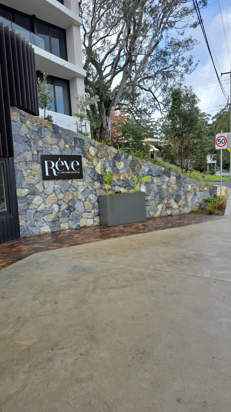 A contemporary entrance features Verona Stone Cladding with a hand-split surface and “Rêve” sign. In front, a large planter with greenery stands beside a 50 km/h speed limit sign and trees. — Stone Cladding