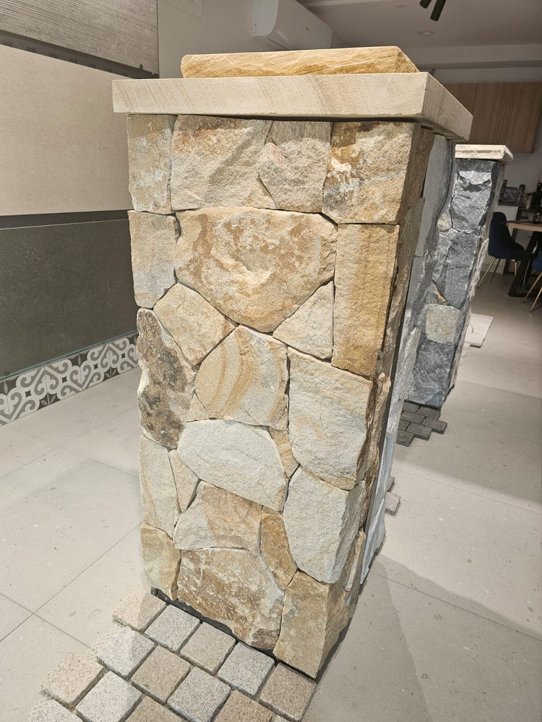 An indoor setting features an Orana Stone Cladding pillar of irregular, light brown and beige natural sandstone with a split surface; stone columns, freestyle walling, and a dining area can be seen in the background. — Stone Cladding