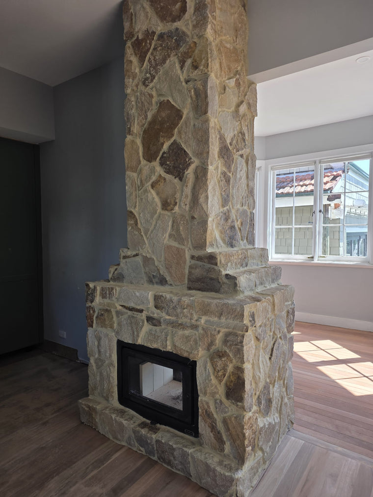 The Orana Stone Cladding fireplace with a glass-fronted firebox stands in a sunlit room with wooden floors and large windows, filling the space with natural light. — Stone Cladding