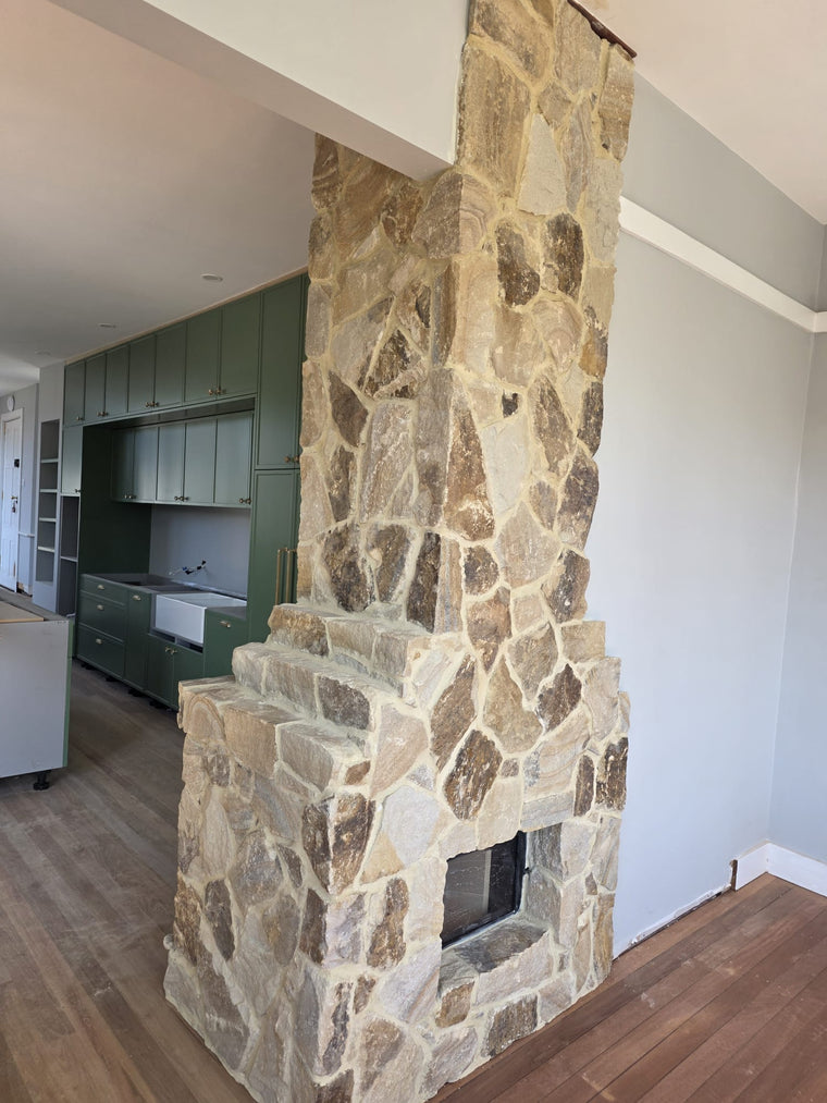 A rustic Orana Stone Cladding fireplace made from natural sandstone and split surface tan-brown stones stands at the center of a room with wooden floors and sage green kitchen cabinets in the background. — Stone Cladding