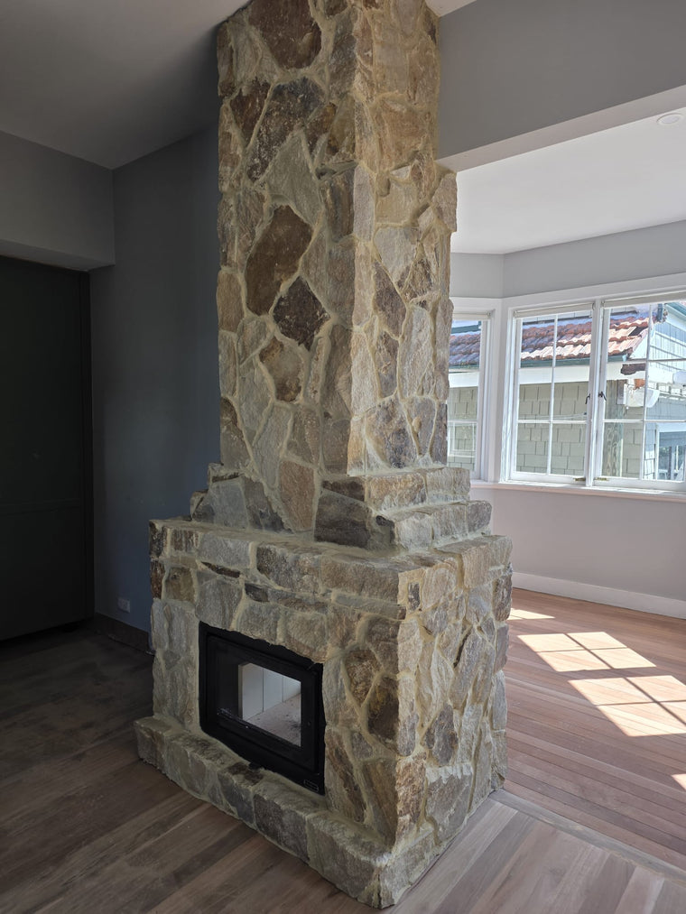 The Orana Stone Cladding with a split surface and glass-fronted firebox is featured in a modern, bright room with large windows and wood flooring, where sunlight highlights the natural sandstone and spacious interior. — Stone Cladding