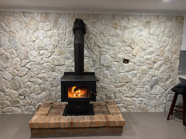 A black wood-burning stove sits on a brick platform against a wall clad in Avelon Stone Cladding. Beautiful free style walling is showcased, with a bar stool and part of a kitchen counter visible on the right.