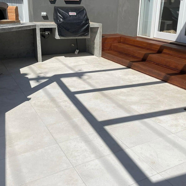 Outdoor patio with Galala Pearl Limestone Pavers, a built-in concrete counter, covered grill, wooden steps, and bold shadow lines from a pergola. Sunlight highlights geometric patterns on the light-toned pavers.