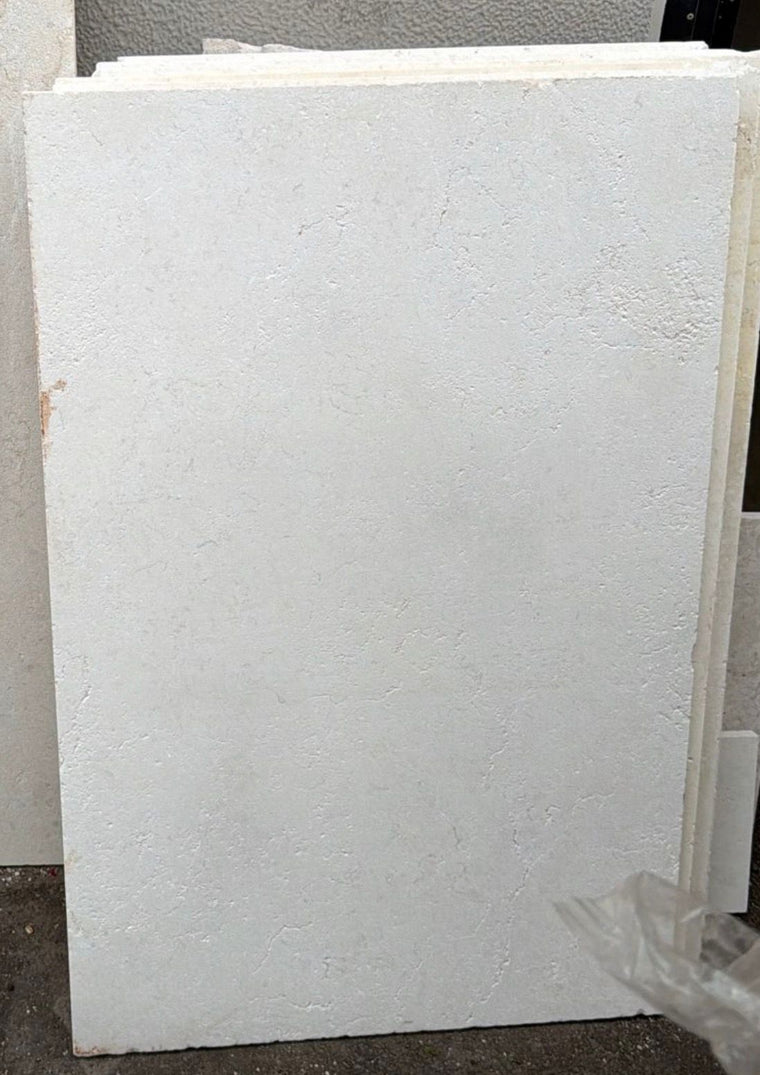 A large, rectangular Galala Pearl Limestone Paver with a subtly textured surface leans against similar tiles on a concrete floor—perfect for wall cladding or pool paving projects. — Natural Stone Paving