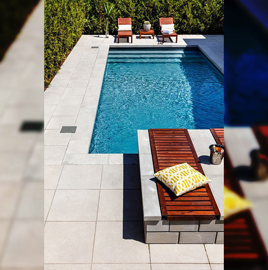 A rectangular backyard pool features Galala Pearl Limestone Pavers, two lounge chairs and a small table at one end, plus a wooden bench with a yellow patterned pillow and metal ice bucket at the other, all surrounded by greenery. — Natural Stone Paving