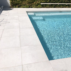 A modern outdoor swimming pool with clear blue water is surrounded by large, light-toned Galala Pearl Limestone Pavers. A metal chair sits on the left, with a green hedge bordering the far side of the pool.