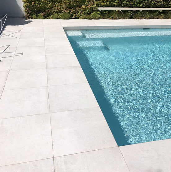 A modern outdoor swimming pool with clear blue water is surrounded by large, light-toned Galala Pearl Limestone Pavers. A metal chair sits on the left, with a green hedge bordering the far side of the pool. — Natural Stone Paving