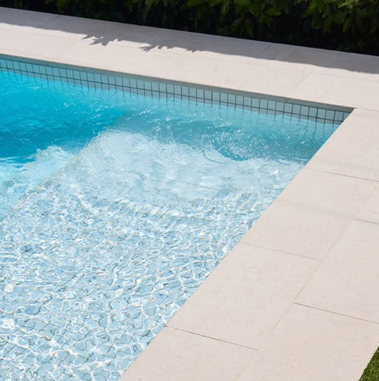 Close-up of a clean swimming pool with turquoise water and light ripples, bordered by Galala Pearl Limestone Pavers and surrounded by lush green foliage in the background. — Natural Stone Paving
