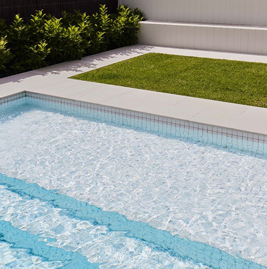 A modern backyard with a clear swimming pool, Galala Pearl Limestone Pavers around the edge, white tile accents, a green lawn patch, and leafy shrubs along a dark fence. — Natural Stone Paving