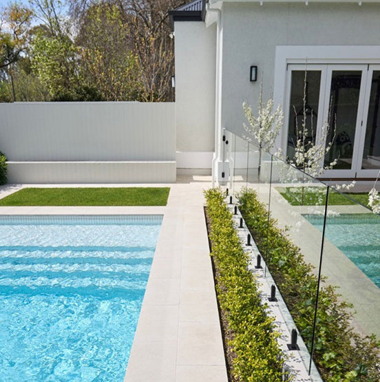 A modern backyard with a clear pool featuring Galala Pearl Limestone Pavers, a glass fence, green hedges, and a white house with large glass doors. The bright area is well-maintained, surrounded by trees and a white privacy fence. — Natural Stone Paving