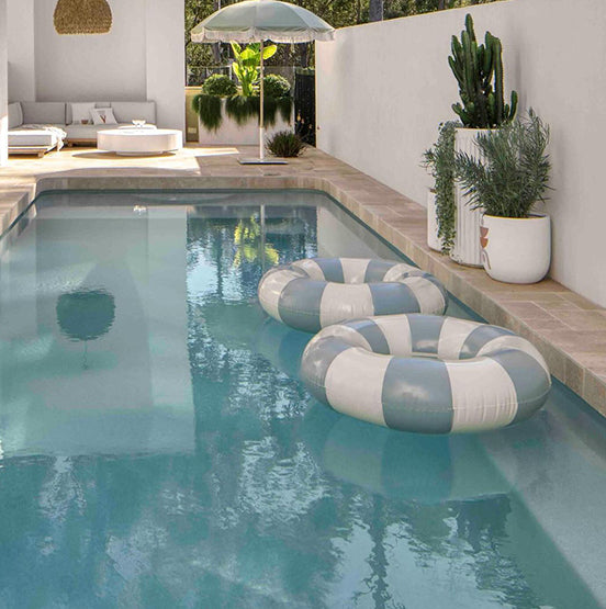 A modern backyard pool features two blue and white striped inflatable rings, potted plants along a white wall with Luxor Cream Limestone Pavers, a large umbrella, lounge seating in the background, and sunlight reflecting on the clear water. — Natural Stone Paving