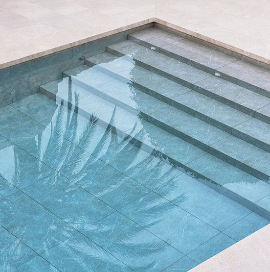 A swimming pool with wide, shallow steps and clear turquoise water reflects palm leaves on the surface, beautifully bordered by Galala Pearl Limestone Pavers. — Natural Stone Paving