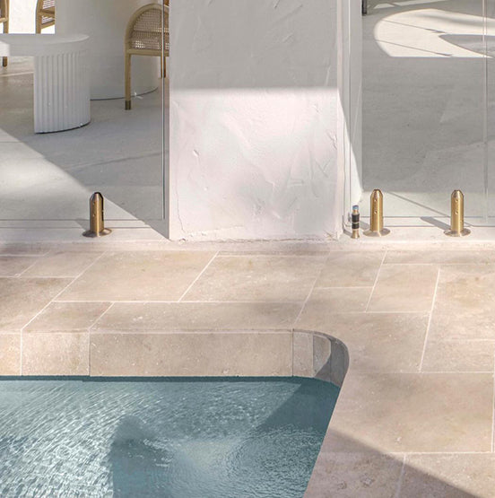 A corner of Luxor Cream Limestone Pavers by a beige tiled patio, with glass doors opening to a modern space with white walls, a white table, and wicker chairs. Sunlight enhances the pavers' beauty for both indoor and outdoor use. — Natural Stone Paving