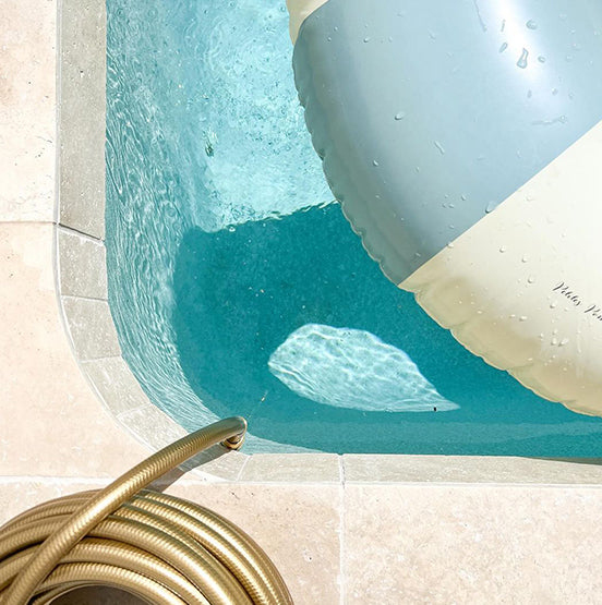 A coiled gold garden hose rests on Luxor Cream Limestone Pavers beside a pool, where a large inflatable float covers part of the water. Sunlight creates shimmering patterns in the clear blue pool, perfect for indoor and outdoor use. — Natural Stone Paving