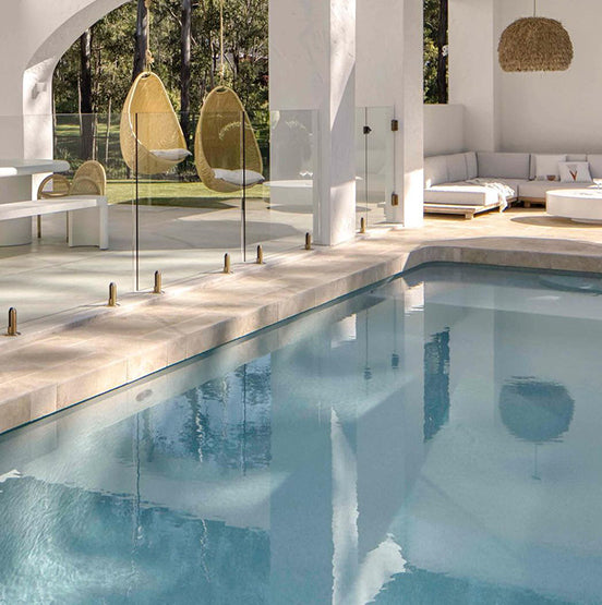 Modern poolside patio features Luxor Cream Limestone Pavers, clear glass railing, white lounge furniture, and two hanging wicker chairs. Large windows showcase green trees. Bright, airy, and stylish—ideal for indoor or outdoor use. — Natural Stone Paving