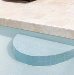 A tiled semicircular pool step leads from a shallow pool into a sunlit area paved with Luxor Cream Limestone Pavers, highlighting their elegance and suitability for indoor or outdoor spaces. A dark outdoor chair sits on the patio above.