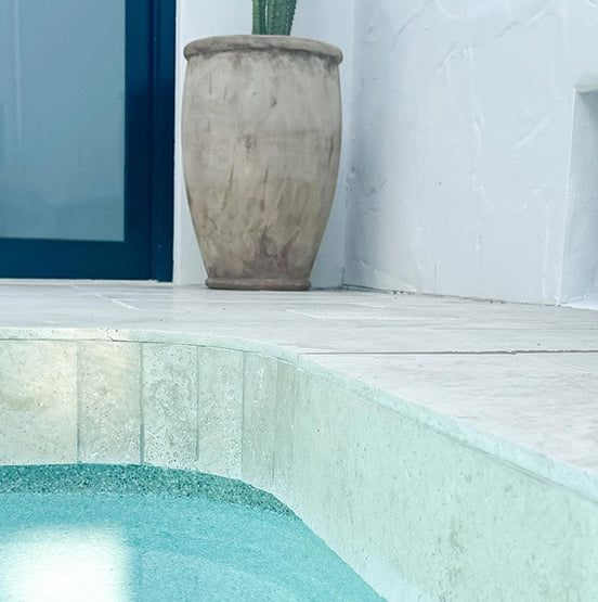A close-up of a pool edge with clear blue water and Luxor Cream Limestone Pavers; a large beige pot with cactus sits by a white textured wall near a blue door—perfect for both indoor and outdoor spaces. — Natural Stone Paving