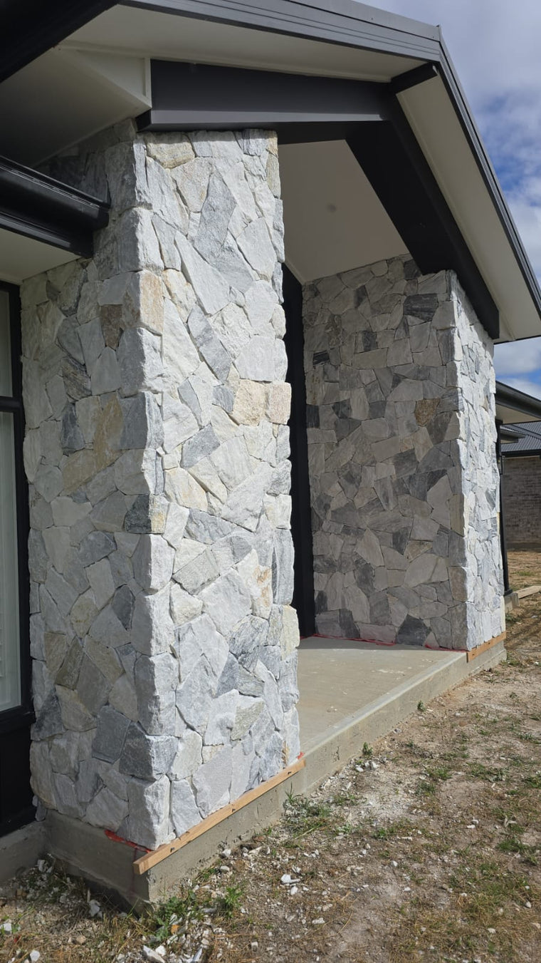 Close-up of a house entrance with two large pillars and a partial wall clad in Luna Grey Stone Cladding (natural quartzite, free style), beneath a modern gabled roof. The concrete porch is bordered by gravel and patches of grass. — Stone Cladding